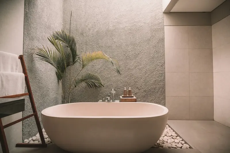 Minimalistisches Badezimmer mit freistehender weisser Badewanne, Kieselsteinen am Boden und Palme vor rauer Betonwand